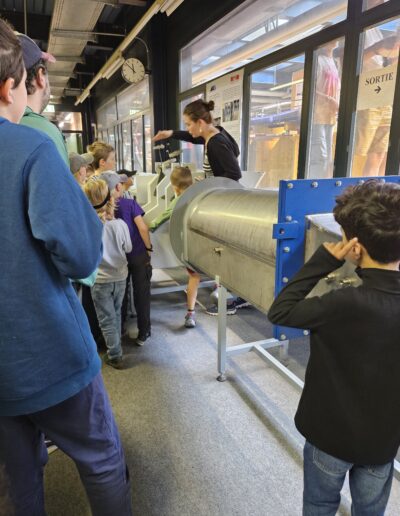 Des enfants et des adultes qui écoutent une femme expliquer à quoi sert une pièce en métal.
