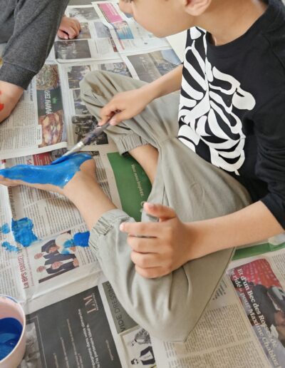 Un enfant assis par terre sur un papier journal qui met de la peinture bleue sous son pied.