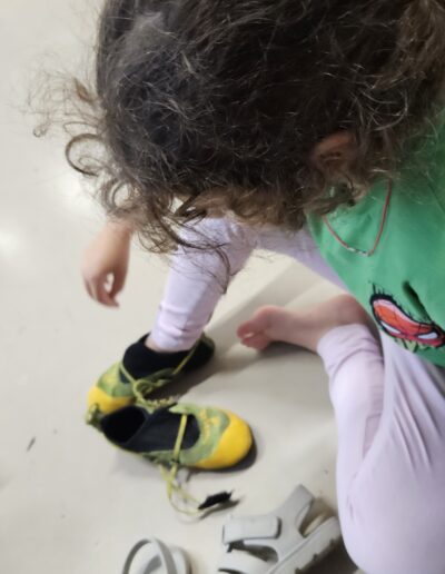 Une petite fille assise par terre qui vient d'enlever ses sandales et enfile un chausson de grimpe.