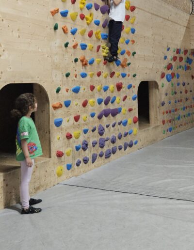 Un mur de grimpe avec des prises avec un petite fille en bas du mur qui regarde un petit garçon qui se trouve au sommet du mur.