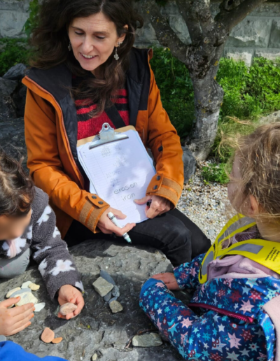 Une adulte assise sous un arbre avec un bloc de feuille à la main qu'elle montre aux trois enfants assis devant elle.