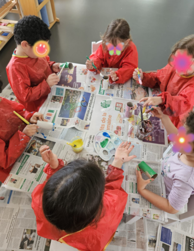Six enfants portant des tabliers oranges assis autour d'une table recouverte de papier journal. Ces enfants font de la peinture.