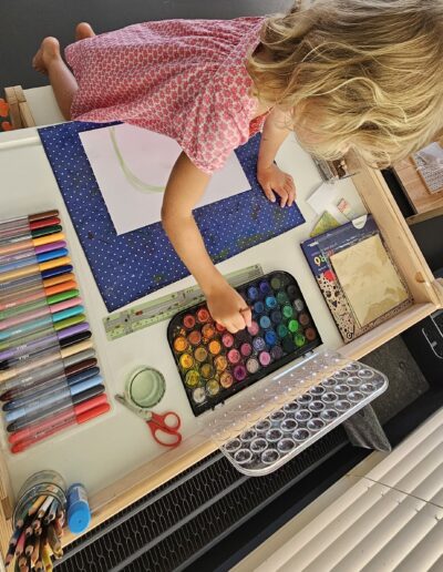 Une petite fille en robe d'été rouge qui peint, elle est en train de mettre de la peinture sur son pinceau.