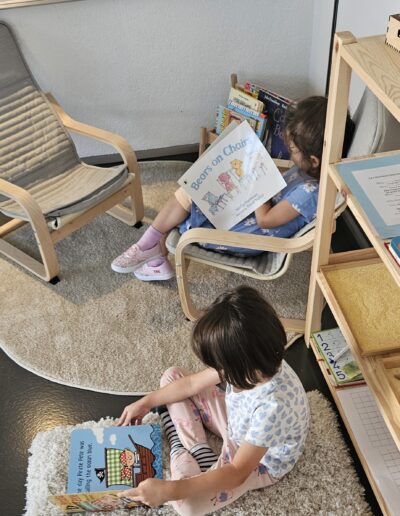 Deux fillettes, une assise dans un fauteuil et l'autre par terre sur un tapis moelleux, qui lisent chacune un livre.