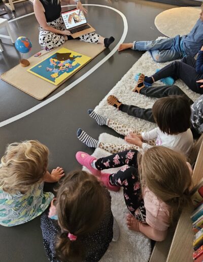 Des enfants assis par terre qui écoute une adulte en train de leur expliquer quelque chose et leur montrer une image sur un ordinateur.