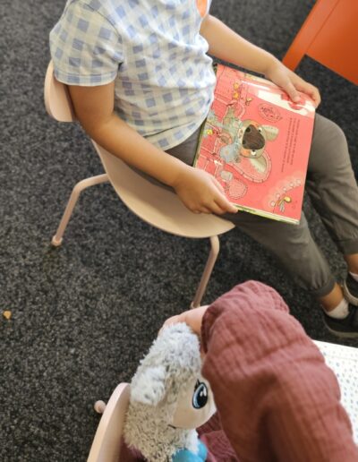 Une enfant assise sur une chaise dans une bibliothèque avec un livre pour enfant ouvert sur les genous.
