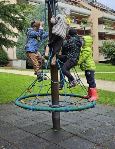 Quatre enfants en habits d'hiver qui sont accrochés à un jeu extérieur. Un poteau autour duquel des cordes sont attachées.