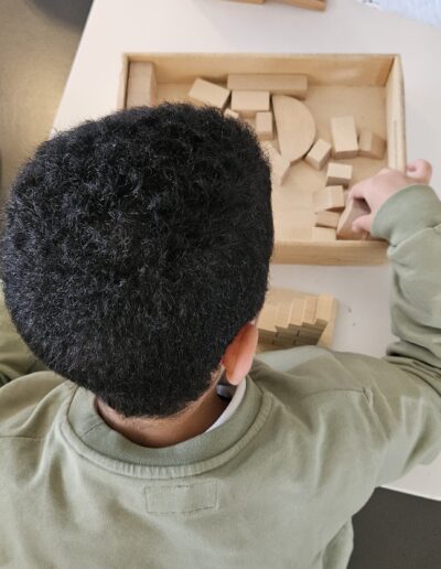 Un petit garçon assis qui joue avec des pièces de construction en bois posées sur une table. En arrière plan, les mains de 2 enfants qui essaient de saisir la même pièce en bois.